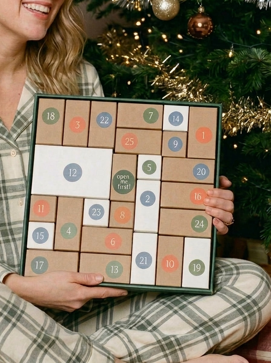 A woman in a plaid pajama shirt holds a Christmas advent calendar in front of a decorated Christmas tree. The game consists of square boxes with numbered compartments in brown, white, blue, green, and orange.
