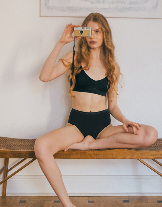 Woman sits cross-legged on a wooden bench against a white wall, holding a vintage-style camera. She is wearing a carbon two-piece set consisting of a scoop-neck bra and high-waisted briefs.