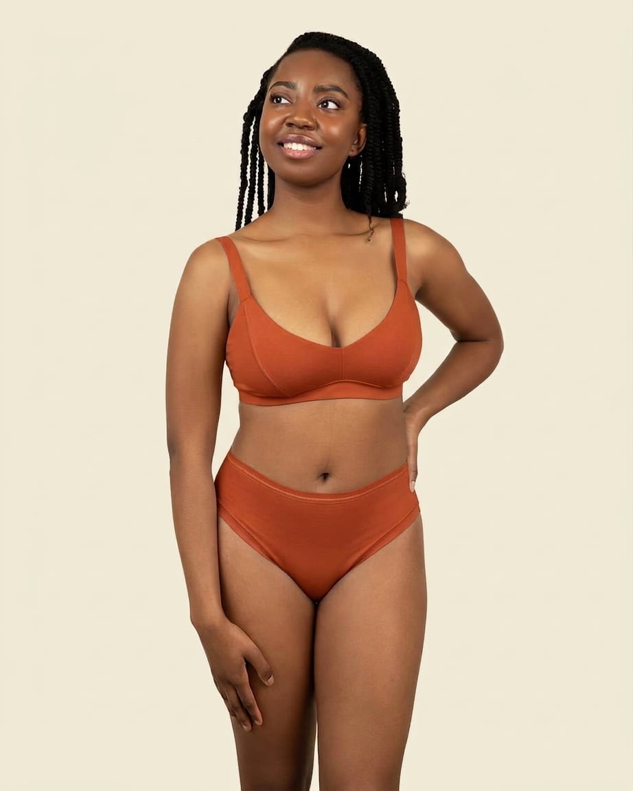 A woman modeling a mid rise bikini in solid terracotta orange color, standing against a plain beige background.