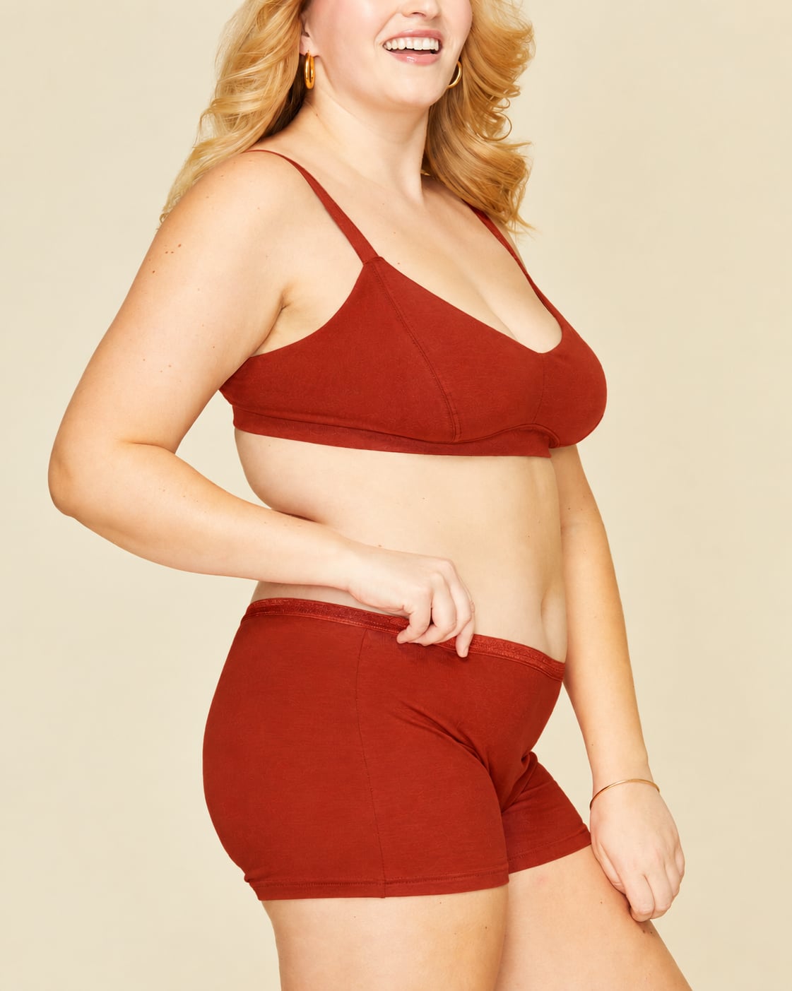A woman modeling mid rise shorties in solid terracotta orange color, standing against a plain beige background.