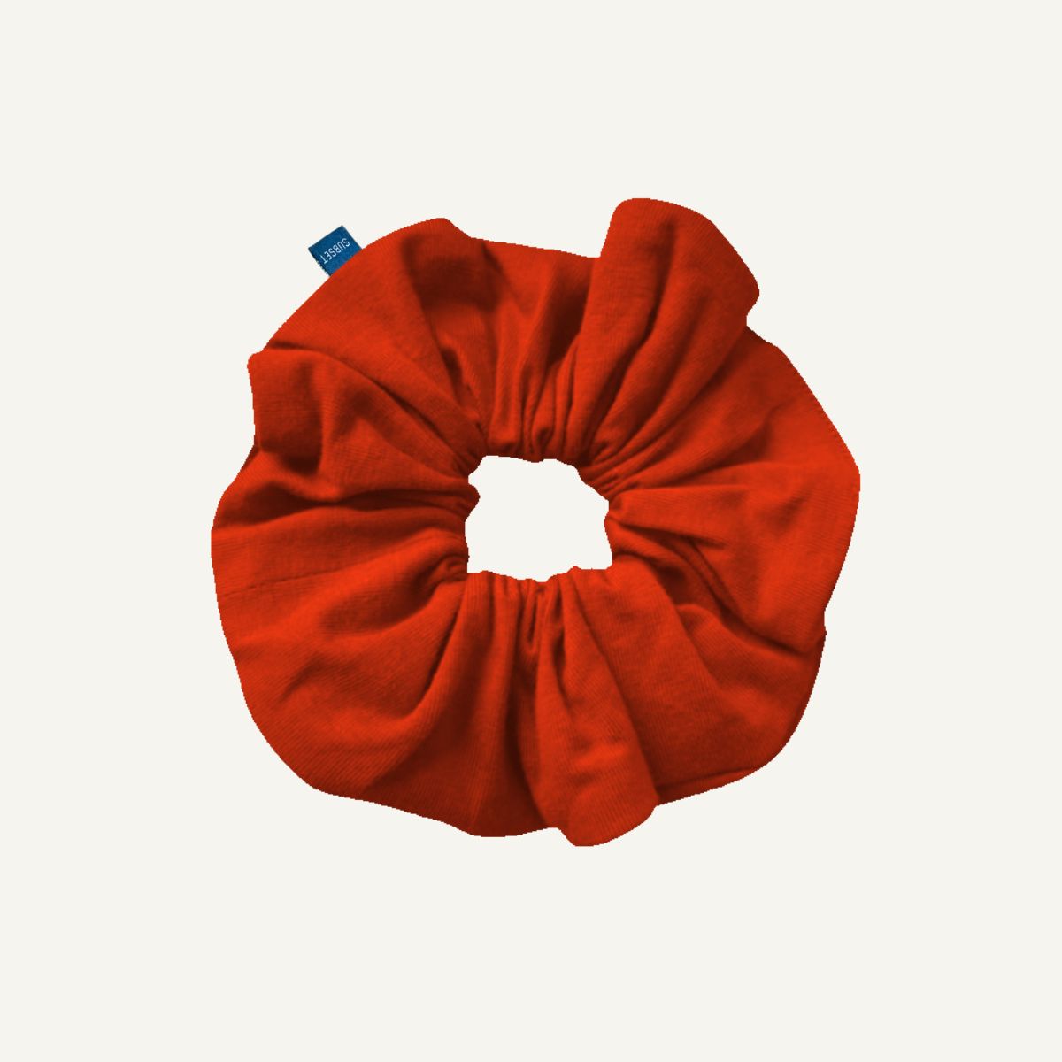 A terracotta orange fabric scrunchie with a small blue brand tag, shown on a plain off-white background.